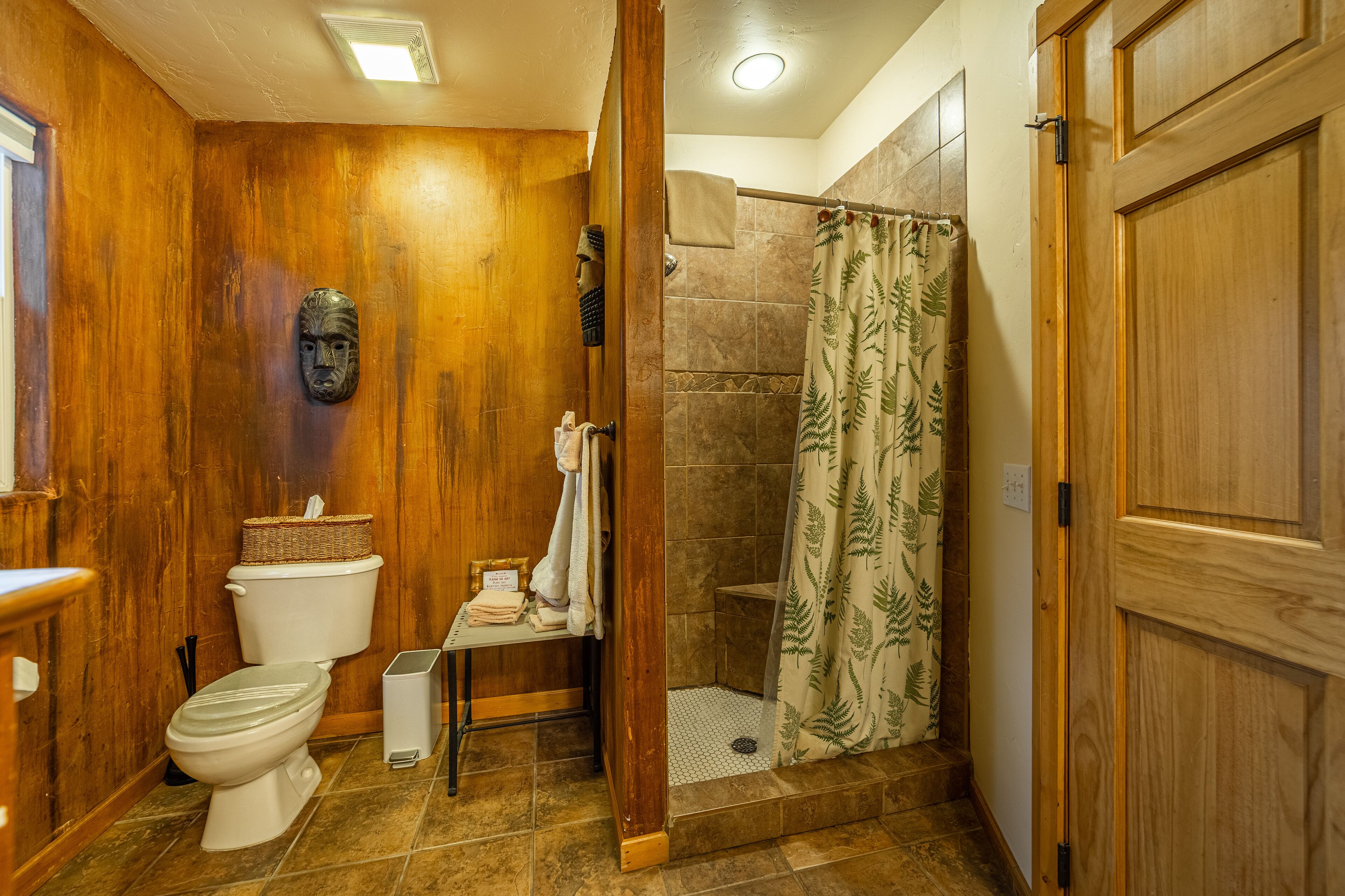A wooden-finished bathroom with a toilet, a shower with a fern-patterned curtain, and a wooden door.