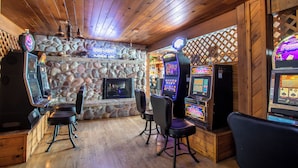 A room with wooden ceiling and stone walls, featuring a stone fireplace, two gaming machines, and three black chairs. - The Islander Inn (Bigfork)