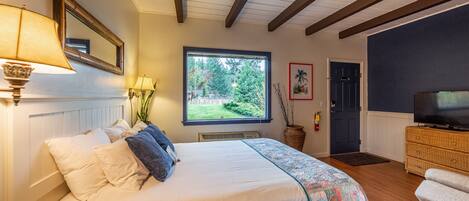 A bedroom with a bed, bedside lamps, a window with a view of greenery, a television on a dresser, and a door.