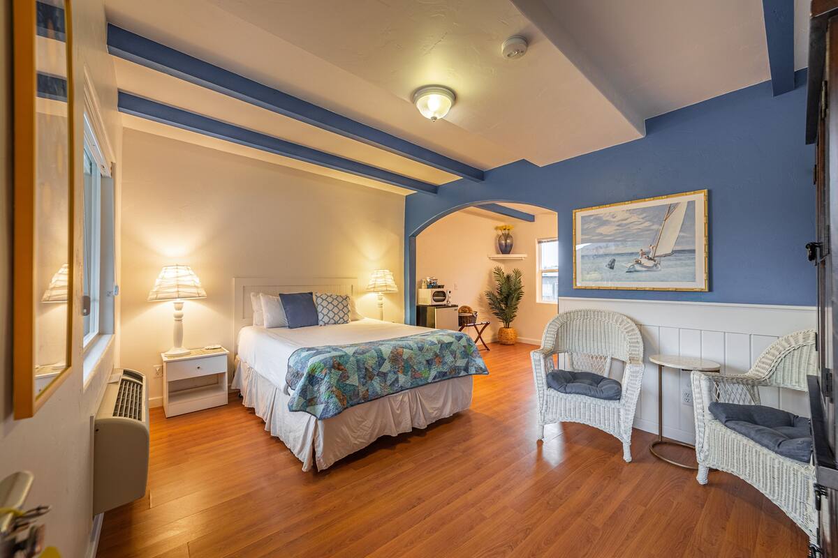 A bedroom with a bed, bedside tables, a chair, a small table, a potted plant, and a painting on the wall.