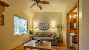 A living room with a wooden floor, a sofa, a coffee table, two wall-mounted lamps, a ceiling fan, and a window with a view of trees. - The Islander Inn (Bigfork)