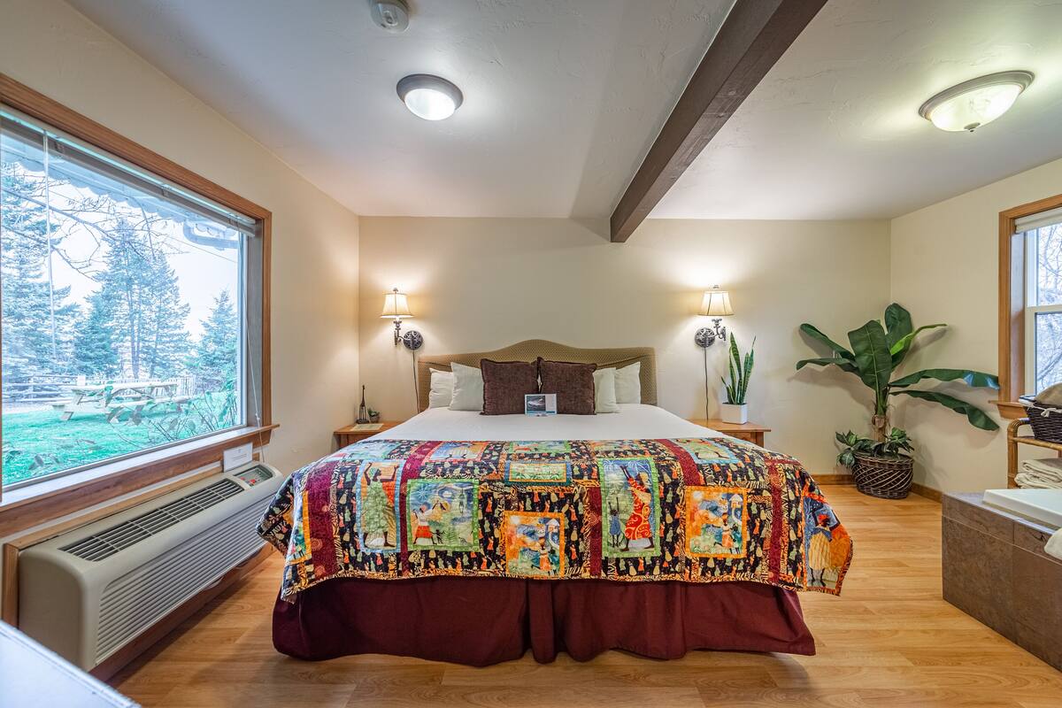 A bedroom with a large bed, two bedside lamps, a window with a view of trees, and a potted plant.