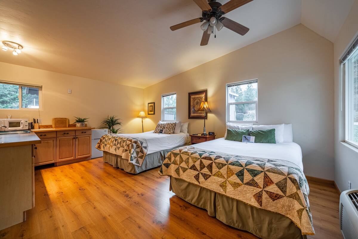 A bedroom with two beds, wooden flooring, a ceiling fan, and a kitchenette.