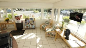 A room with a tiled floor, a wooden table with a computer, a magazine rack, two cushioned chairs, a television, and a window with a view of the outside. - Victoria Motel (Ventura)