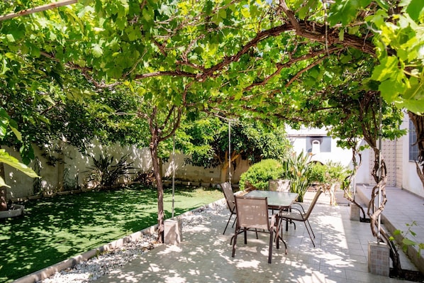 Une terrasse avec une table et des chaises, entourée de verdure et avec un bâtiment blanc en arrière-plan.