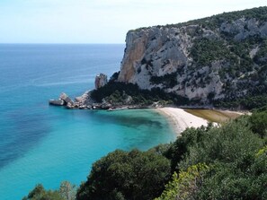 Paesaggio costiero con spiaggia, acque cristalline turchesi e una scogliera rocciosa.