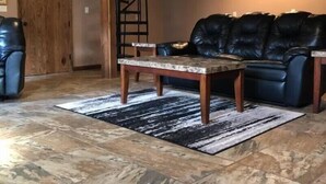 A living room with a stone tile floor, a black leather sofa, a wooden coffee table, and a black and white rug. - Elk from the Hot Tub - Bugle Inn (Weedville)