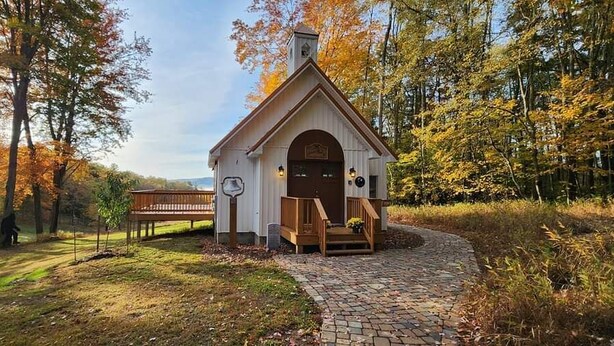 Una pequeña capilla con una puerta de madera y escalones que llevan a ella, rodeada de árboles y un sendero de ladrillo.