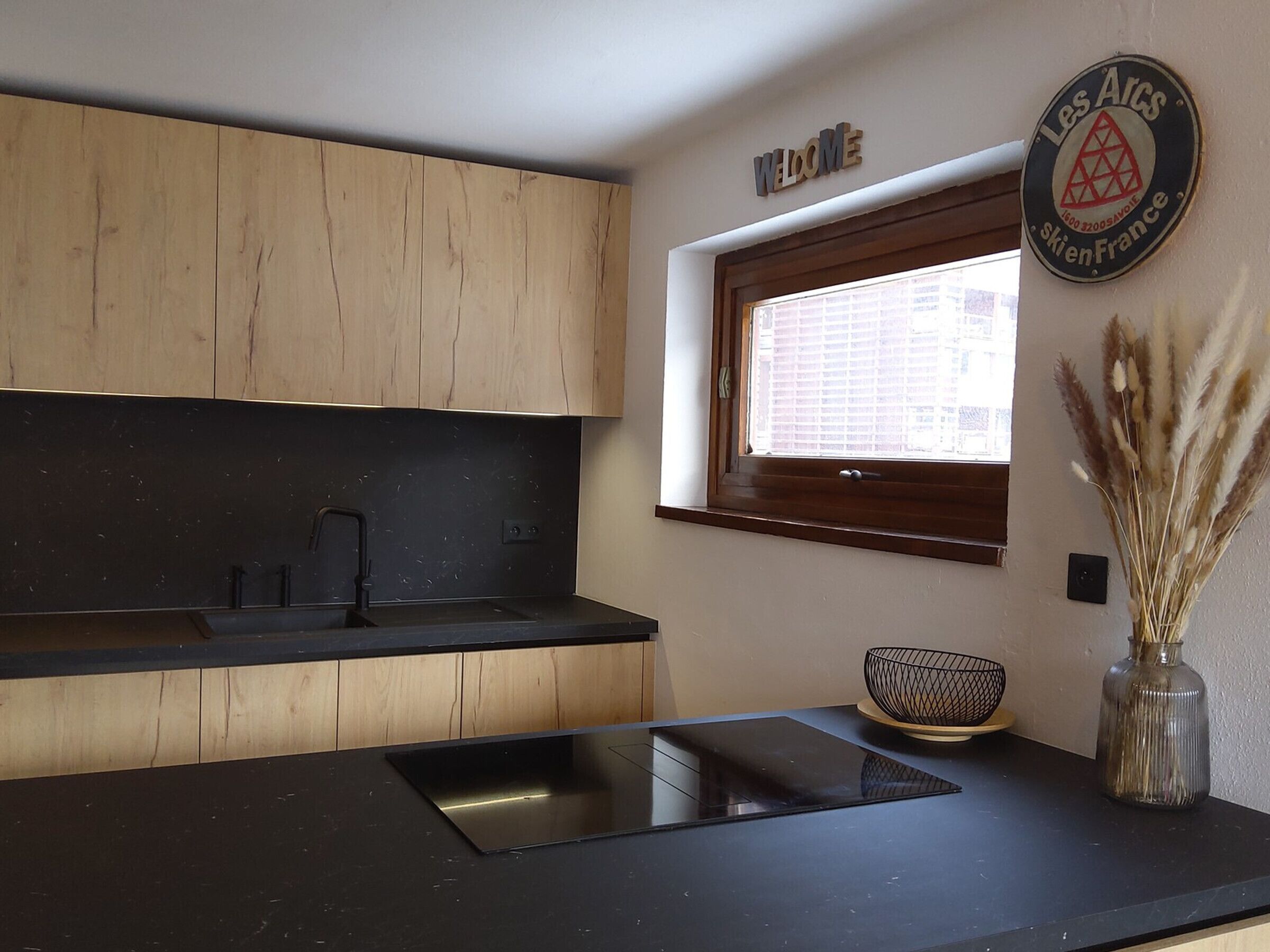 A modern kitchen with wooden cabinets, a black countertop, a window with blinds, a welcome sign, and a decorative vase.