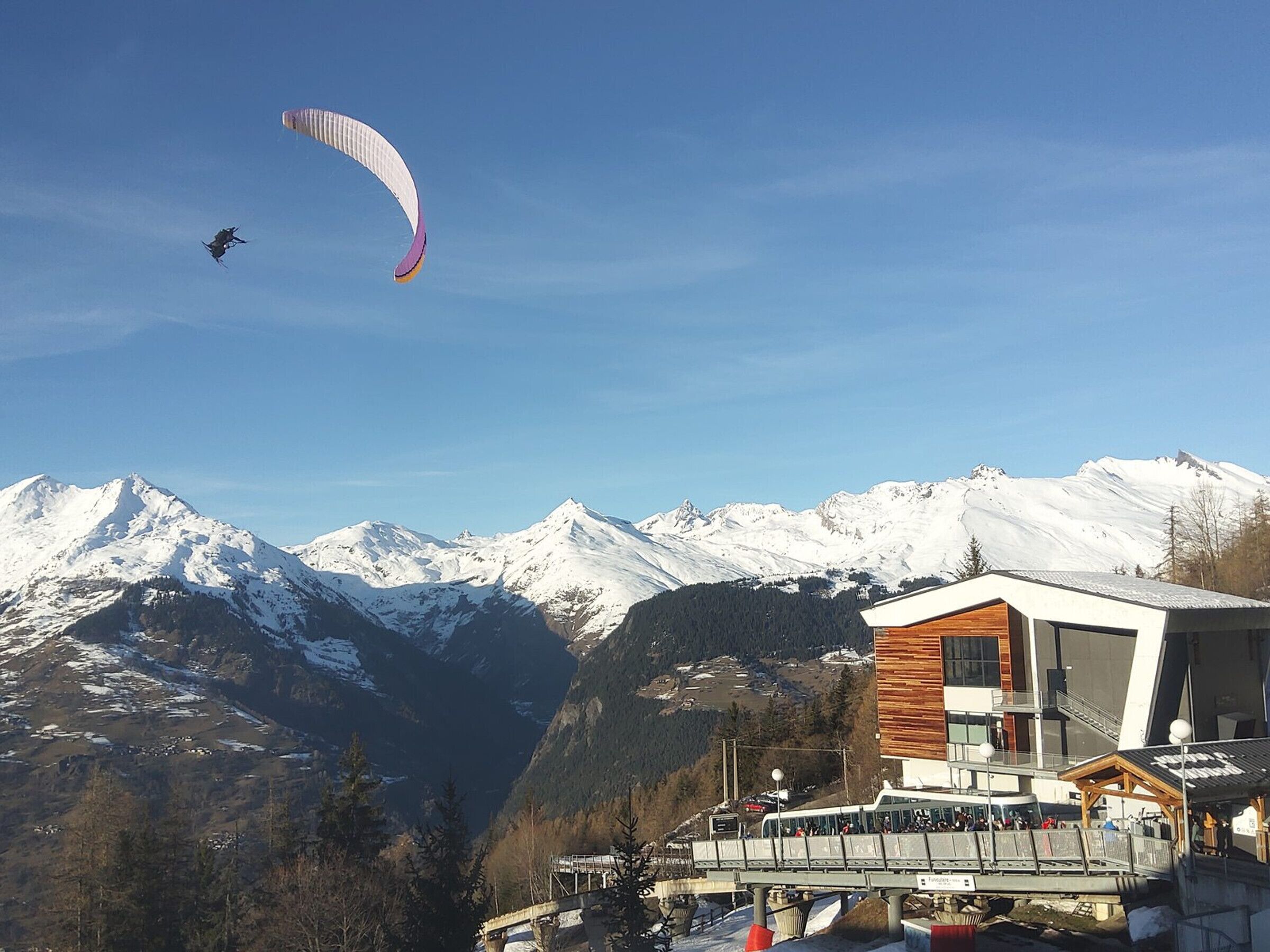 Een bergresort met een modern gebouw, een skilift en iemand die paraglide.'