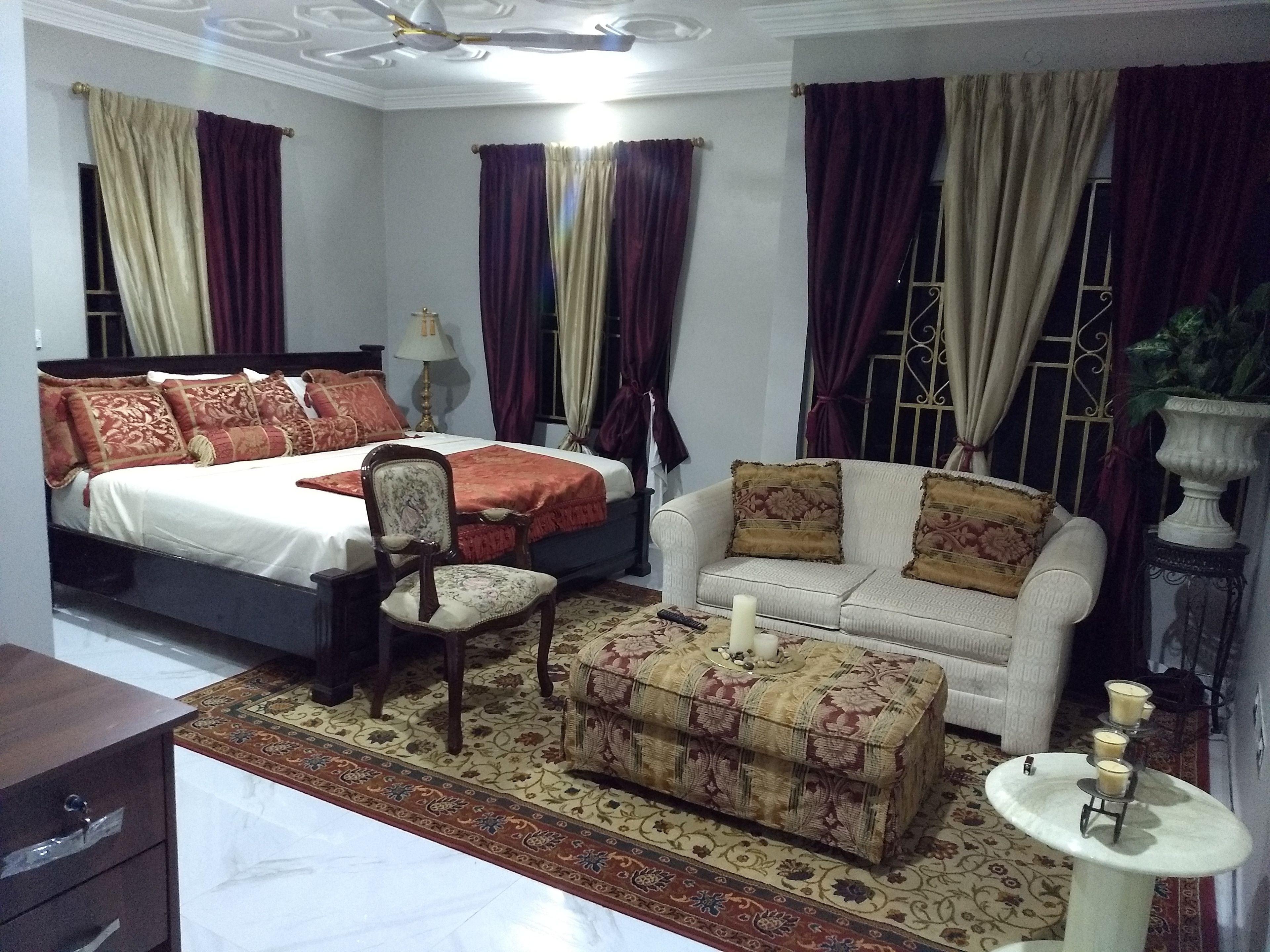 A bedroom with a bed, a sofa, a chair, a small table with candles, and a decorative plant.