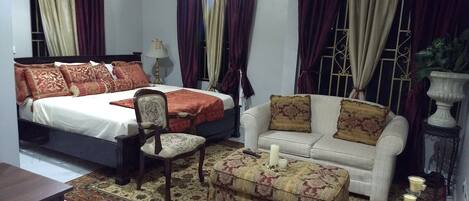 A bedroom with a bed, a sofa, a chair, a small table with candles, and a decorative plant.