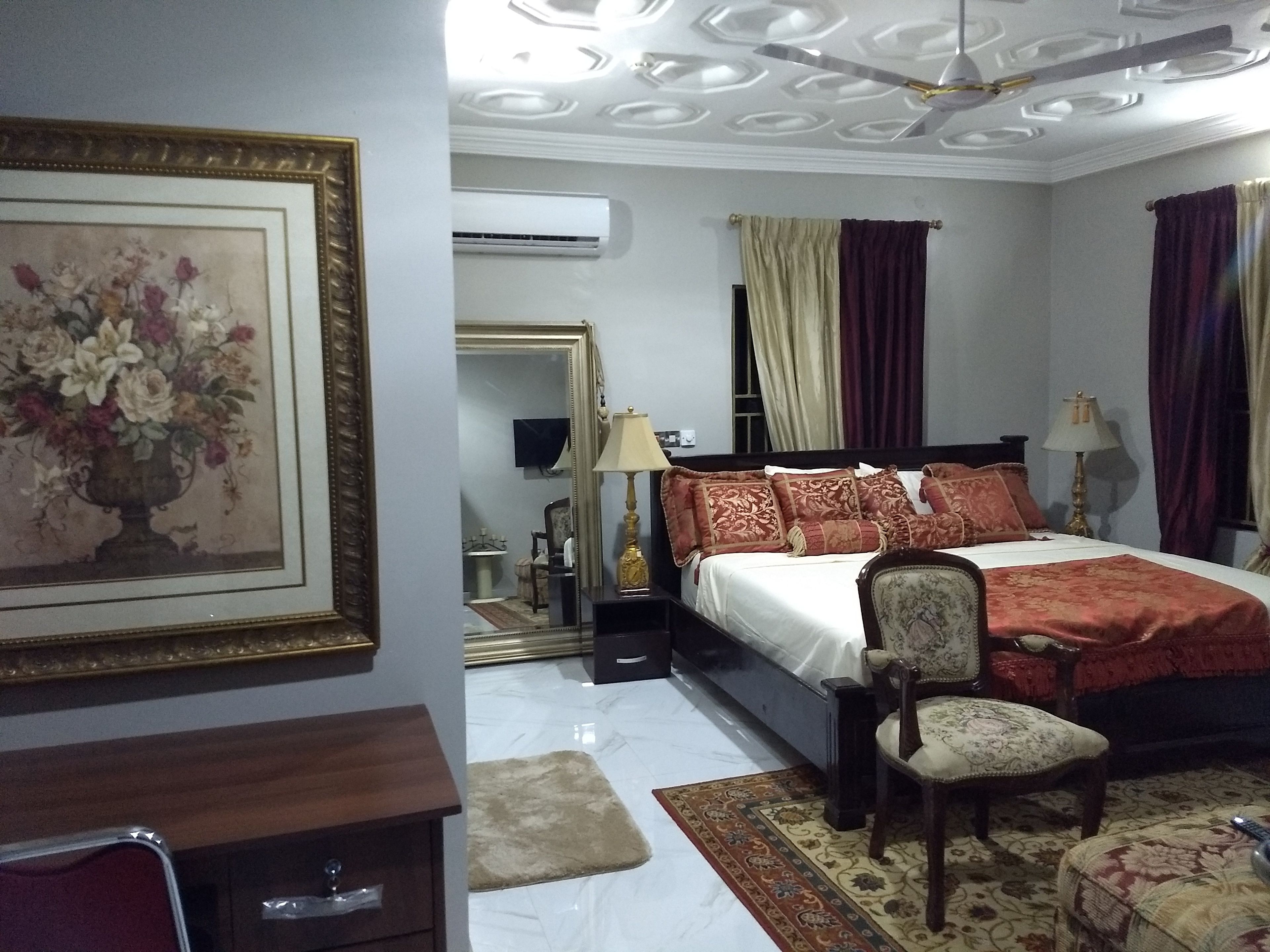 A hotel room with a bed, a chair, a nightstand, a lamp, a mirror, and a framed picture on the wall.