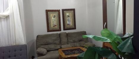 A living room with a sofa, a coffee table, a chair, and a large plant. There are two framed pictures on the wall.