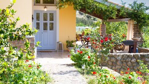 Un bâtiment jaune avec une porte blanche, un mur en pierre et un jardin où poussent des fleurs rouges.