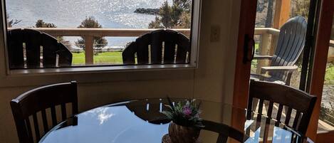 A dining area with a glass table and chairs, a view of a lake and mountains, and a wooden deck outside.