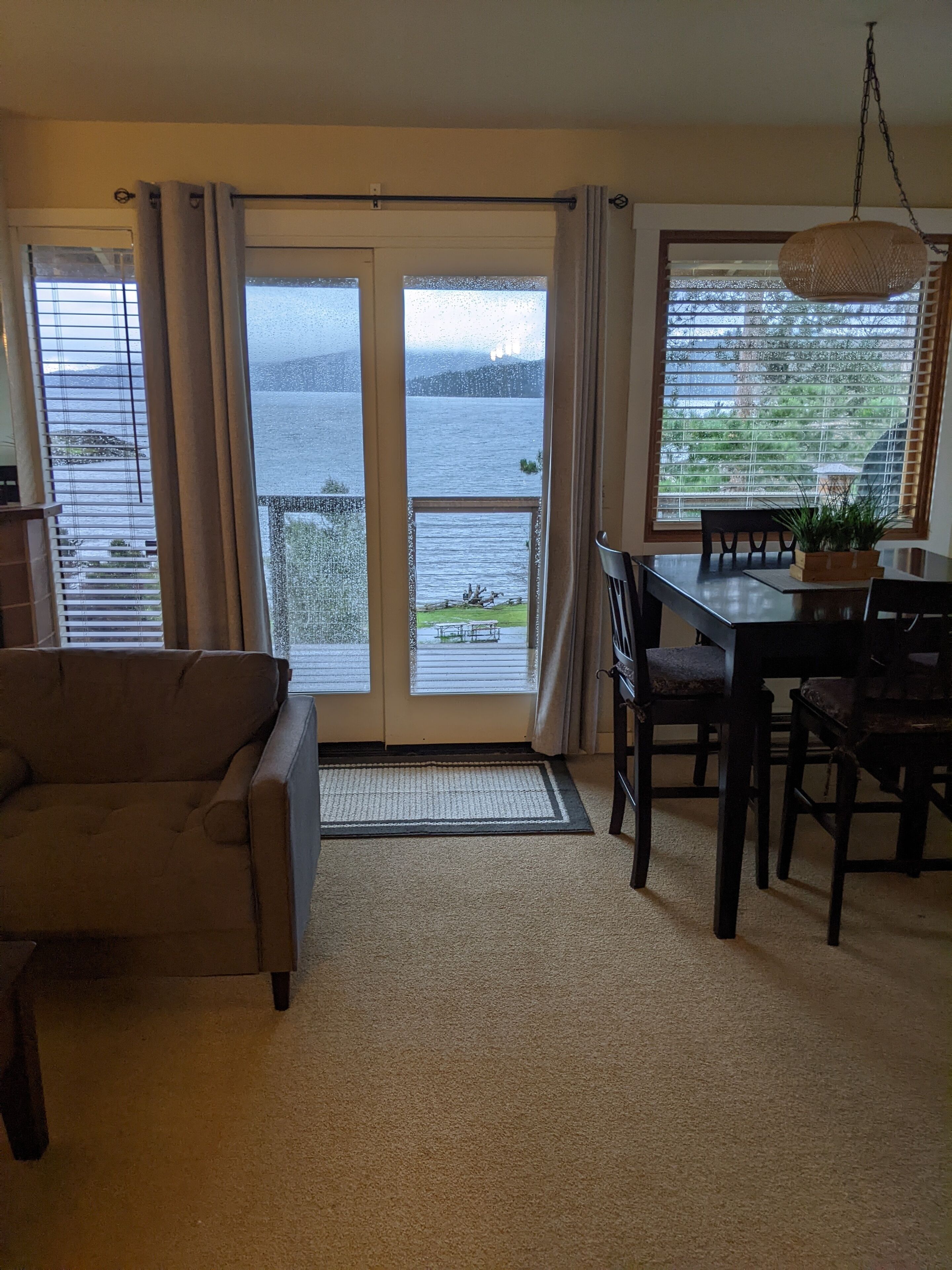 A room with a sofa, a dining table with chairs, a potted plant, and a view of a body of water through large windows.