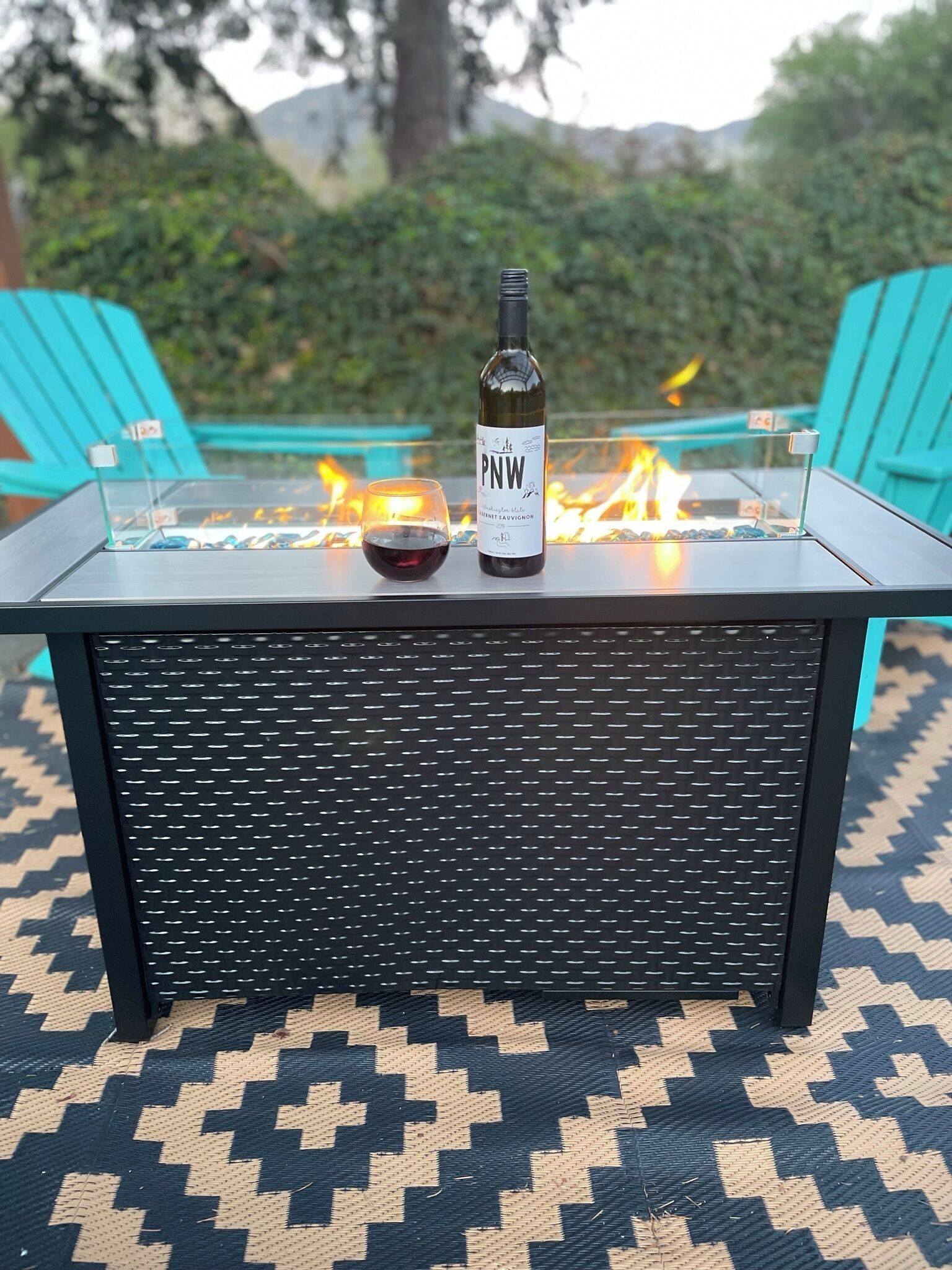 Uma mesa preta para áreas externas com uma garrafa de vinho e um copo em cima, cercada por um elemento de fogo e cadeiras azul-turquesa.