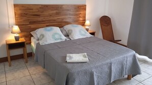 A neatly made bed with a grey bedspread and two pillows, a wooden headboard, two bedside lamps, and a wooden chair. - La Rivière St Louis, 2-room apartment "Furnished tourist accommodation 3 *** (Le Ouaki)
