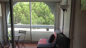 A balcony with a table and chairs, a bed with a TV, and a view of trees through a glass door. - Studio Pool Parking (Le Grau-du-Roi)