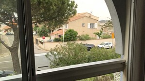 A view from a balcony with a window, overlooking a residential street with houses, parked cars, and greenery. - Studio Pool Parking (Le Grau-du-Roi)