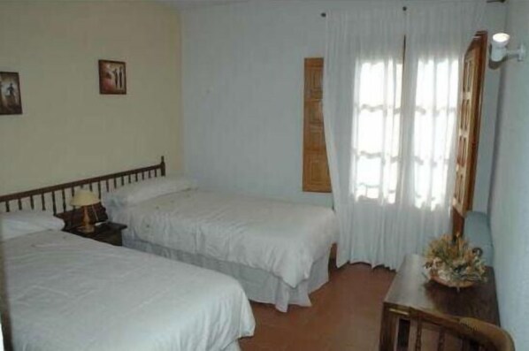 A room with two single beds, a wooden nightstand with a lamp, a wooden chair, and a small table with a vase.