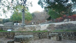 A stone structure with steps leading up to a central pillar, surrounded by trees and a stone wall.