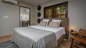 Een slaapkamer met een bed, nachtkastje, stoel, raam met uitzicht en een badkamer zichtbaar door een geopende deur.