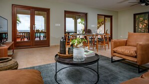 Een ruime woonkamer met een grote schuifpui naar buiten, een comfortabele leren bank, een salontafel met decoratieve objecten, en een televisie op een houten dressoir.