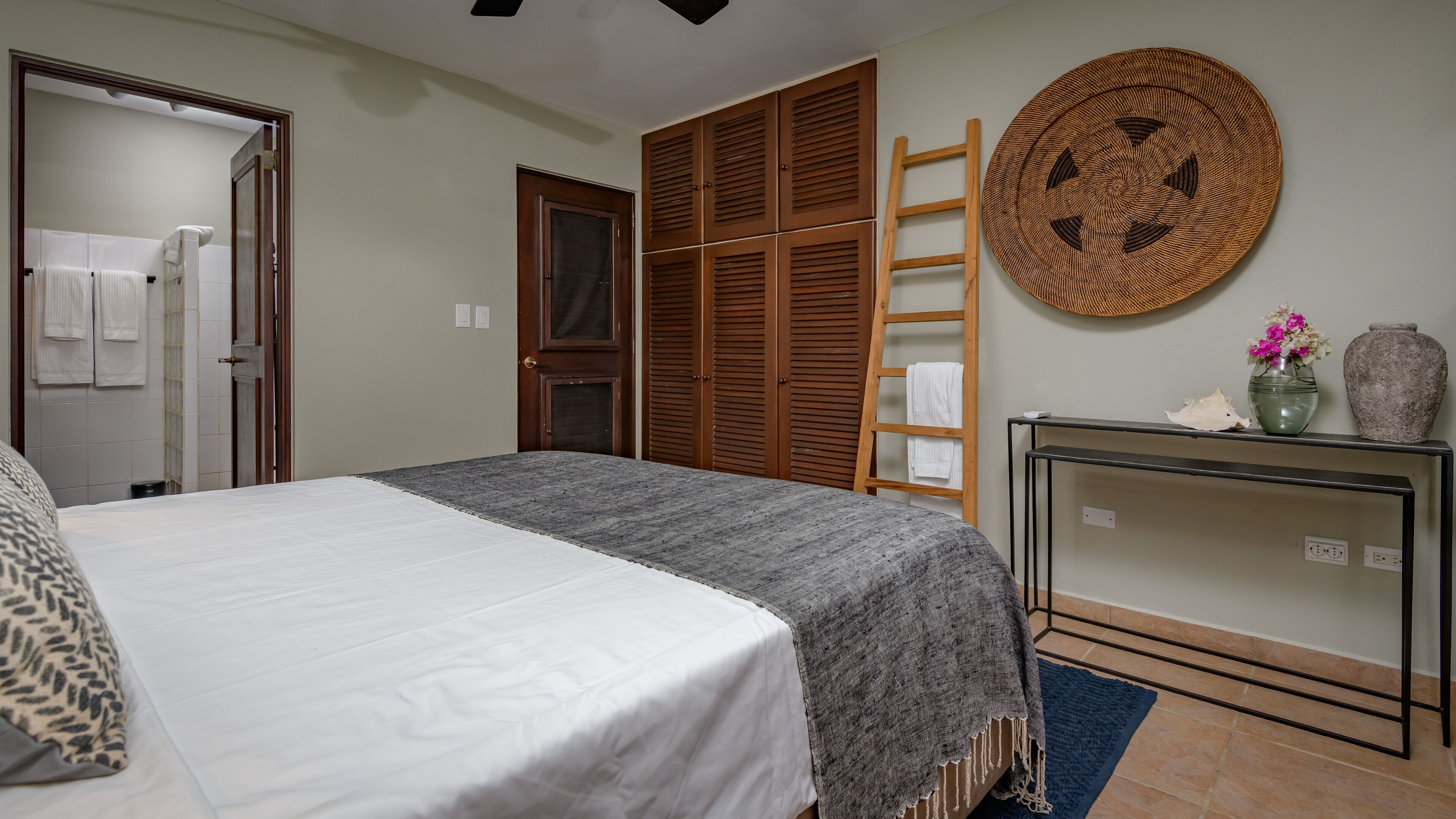 Un dormitorio ordenado con cama, armario, mesita de noche con un florero y un adorno circular en la pared.