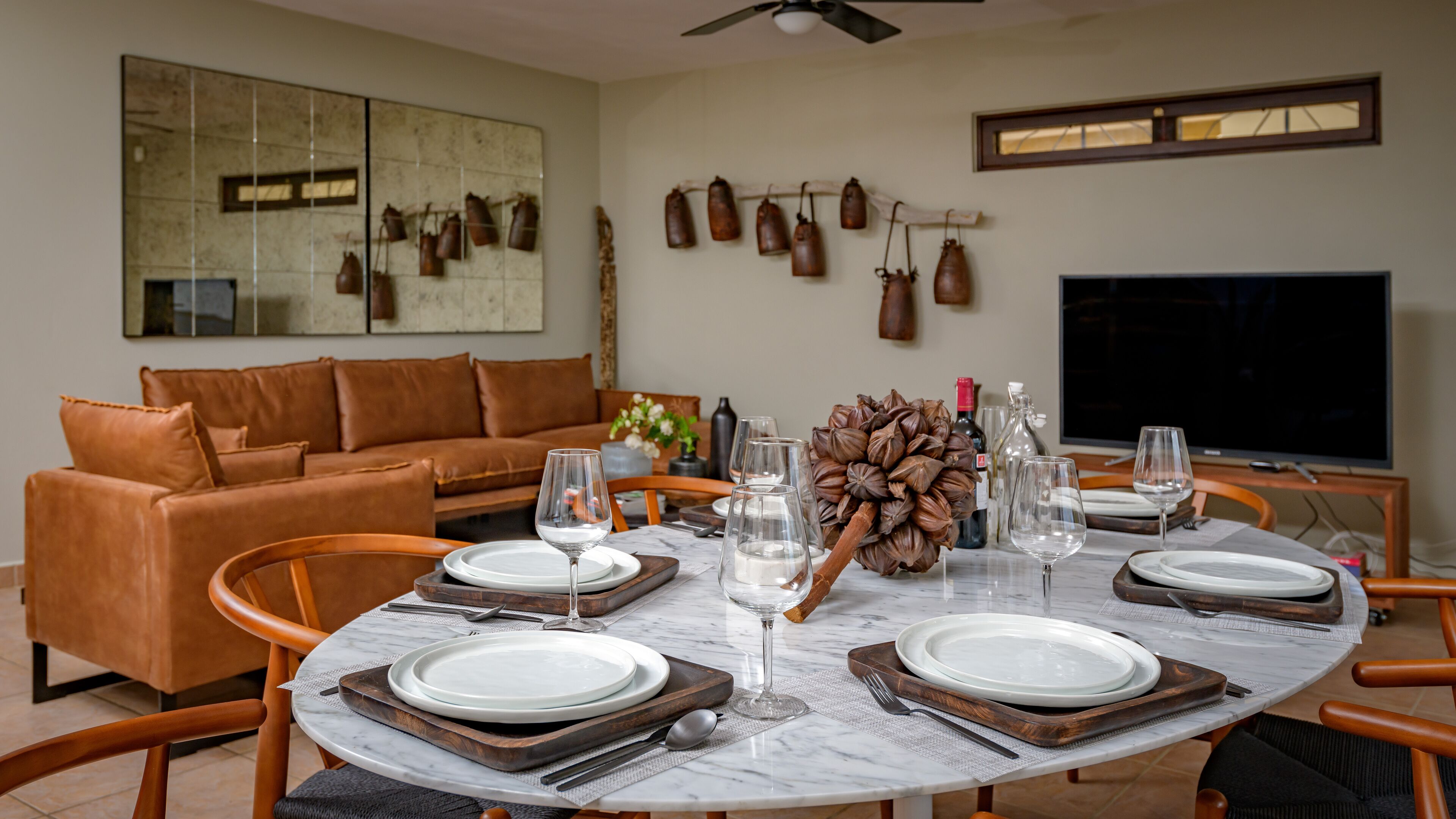 Uma sala de jantar com uma mesa redonda posta para quatro pessoas, rodeada por sofás de couro e um espelho grande.