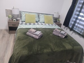 Un lit bien fait, recouvert d’une couverture verte, avec deux oreillers et des serviettes pliées posées sur une table de chevet en bois.