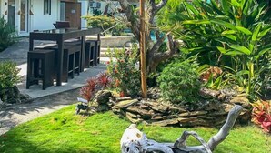 Un jardín tropical con un área de comedor, una gran escultura de árbol y una casa con techo verde.