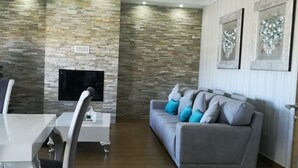 A modern living room with a stone wall, a fireplace, a grey sofa with blue cushions, and a dining area with a white table and chairs. - HOUSE WITH SWIMMING POOL AND BARBECUE (Vila Boim)