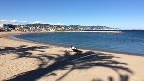 A beach with a person sitting on a blanket, a clear sky, and a distant cityscape. - ADORABLE T1 ALCOVE RESIDENCE SECURE WITH 3 POOLS (Mandelieu-la-Napoule)