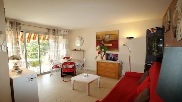 A living room with a red sofa, a white coffee table, and a dining area with a table and chairs. There is a large window with curtains, a floor lamp, and a painting on the wall. - ADORABLE T1 ALCOVE RESIDENCE SECURE WITH 3 POOLS (Mandelieu-la-Napoule)