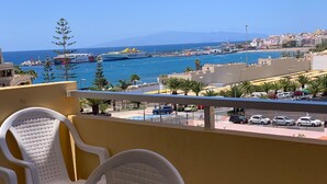 A balcony with white plastic chairs and a table, overlooking a coastal area with a marina, parked cars, and buildings. - Views of Los Cristianos pier. Pier View. (LOS CRISTIANOS)