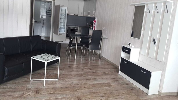 Una sala de estar moderna con un sofá negro, una mesita blanca pequeña y un comedor con una mesa negra y sillas. Hay una zona de cocina con armarios y un refrigerador.