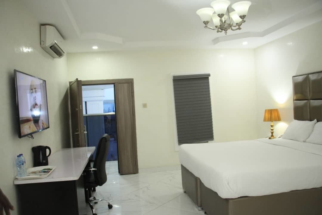 A hotel room with a bed, a desk with a chair, a television, a lamp, and a window with blinds.