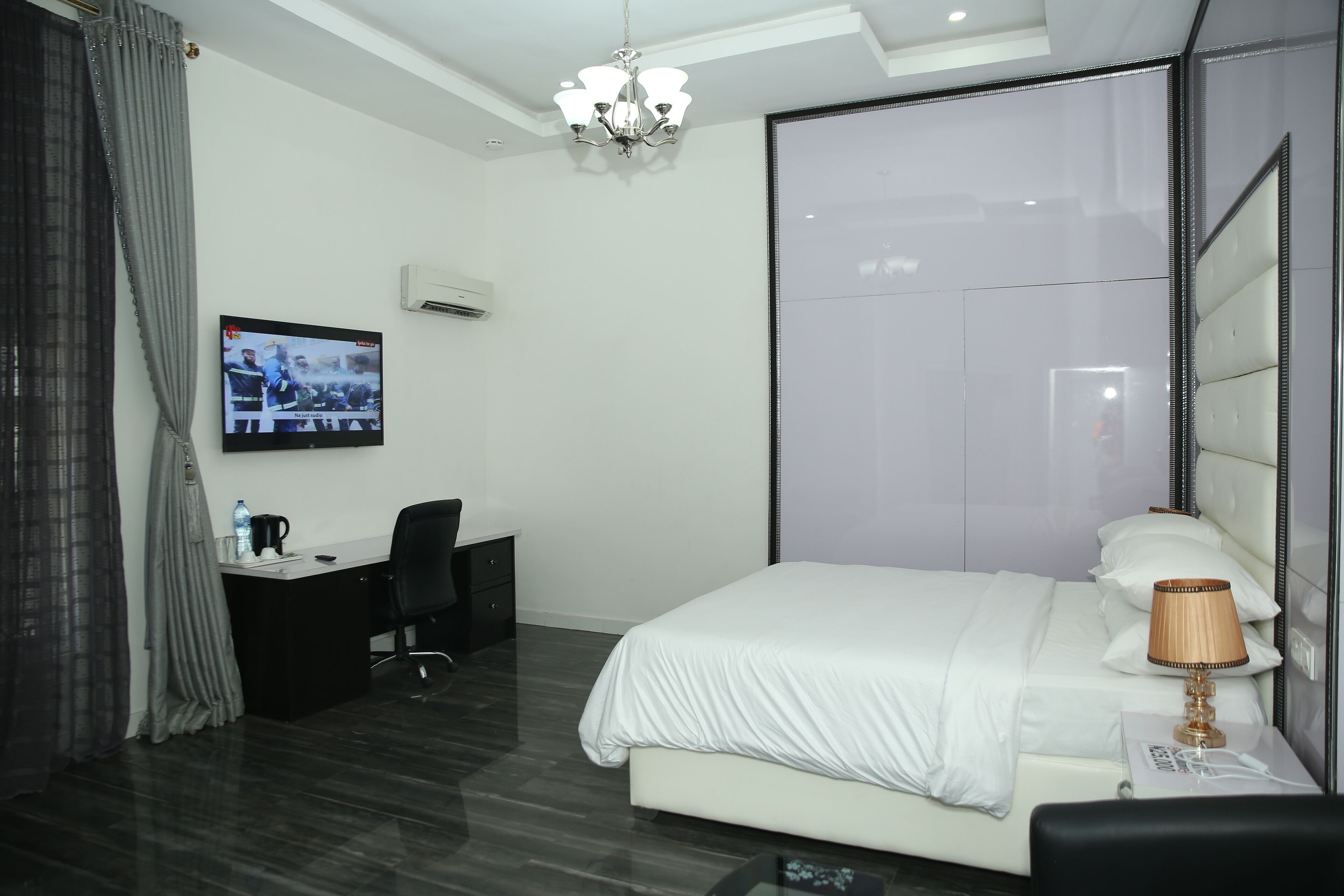 A modern hotel room with a large bed, a desk with a chair, a television mounted on the wall, and a chandelier.