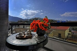 Un balcón con una mesa lista para el té, un jarrón de flores y vistas a edificios y montañas.