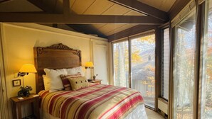 Un dormitorio con una cama grande, dos lámparas de noche, una mesita de noche y vistas a árboles otoñales a través de amplias ventanas.