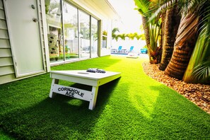 A patio with artificial grass, a white table labeled 'Cornhole Age,' and a pool area with lounge chairs.