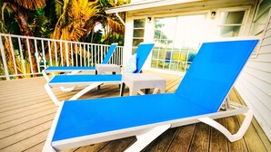 Zwei blaue Lounge-Stühle auf einer Holzterrasse mit weißer Geländer und Blick auf Palmen.