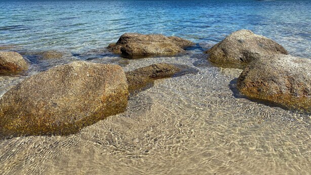 A clear coastal view with large rocks in shallow water, a distant shoreline with buildings, and a sailboat.