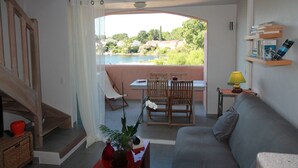 A living room with a sofa, a dining table with chairs, a small table with a lamp, and a view of a lake and houses.