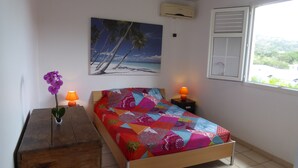 Ein Schlafzimmer mit farbenfroher gesteppter Bettdecke, einem Holznachttisch mit einer Vase voller lila Blumen, zwei Wandleuchten, einem Strand-Motivbild über dem Bett und einem Fenster mit Blick auf Grünflächen.