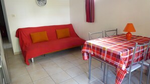 Ein Zimmer mit einem roten Sofa, zwei orangenen Kissen, einem Metalltisch mit kariertem Tischläufer und einer orangenen Tischlampe.