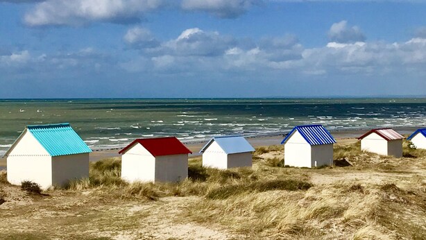 Une rangée de cabanes de plage aux toits bleus et rouges, situées sur une plage de sable, avec l’océan en arrière-plan.