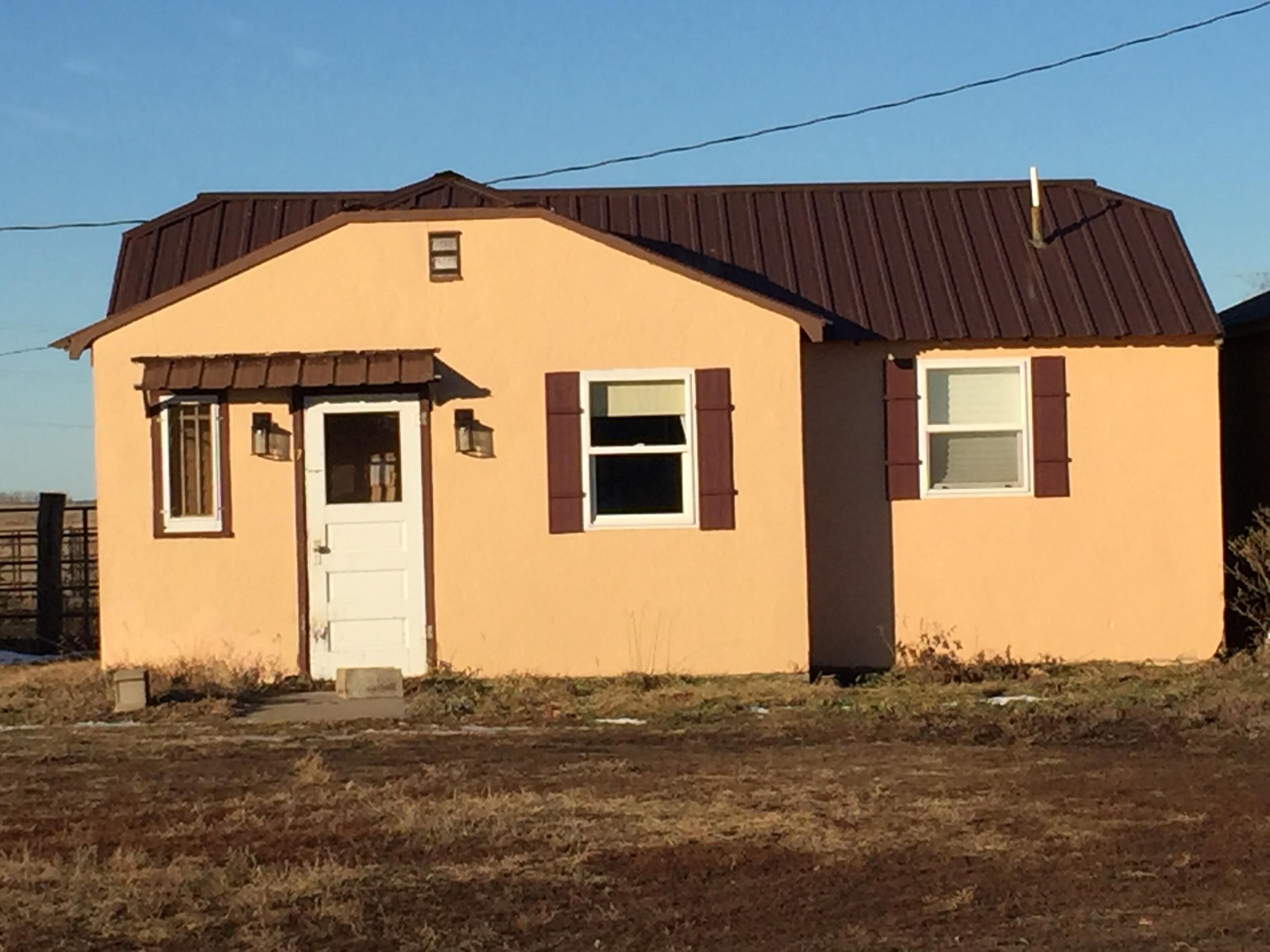 A single-story house with a brown roof and beige walls, featuring a front door, two windows with shutters, and a small porch area.